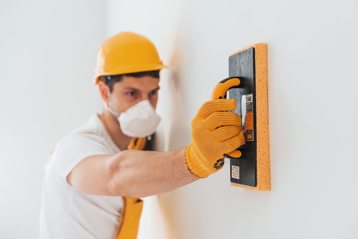 Handyman in yellow uniform and protective mask polishing wall indoors. House renovation conception.
